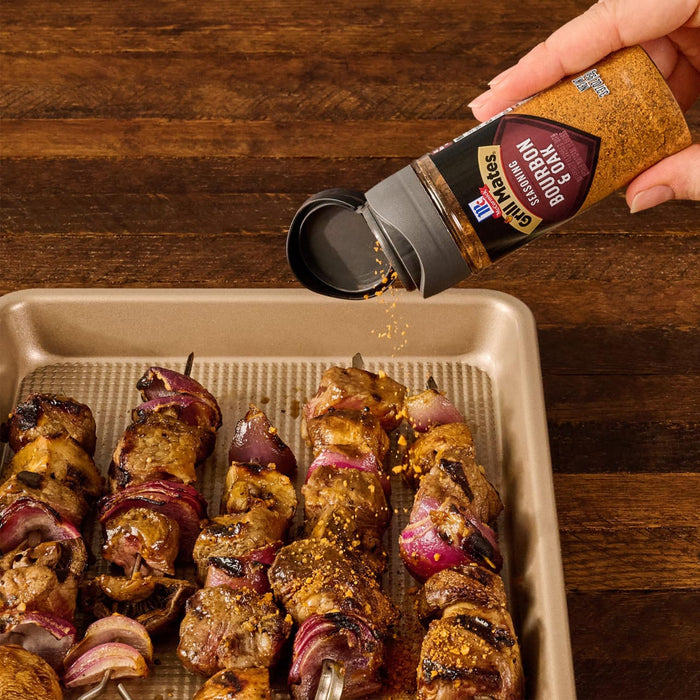 A hand sprinkles Mccormick Grill Mates Bourbon &amp; Oak Seasoning Ground 3.37 Oz over freshly grilled meat and vegetable skewers on a baking sheet.