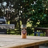 A bottle of Mccormick Grill Mates Bourbon &amp; Oak Seasoning Ground 3.37 Oz sits on a wooden picnic table in a backyard, with a grill and trees with string lights blurred in the background.