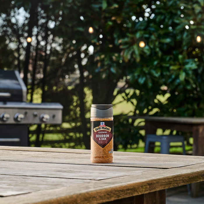 A bottle of Mccormick Grill Mates Bourbon &amp; Oak Seasoning Ground 3.37 Oz sits on a wooden picnic table in a backyard, with a grill and trees with string lights blurred in the background.