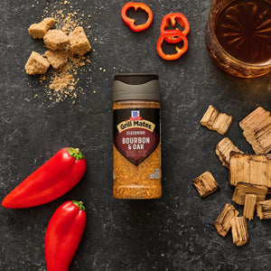 A bottle of McCormick Grill Mates Bourbon &amp; Oak Seasoning is displayed with brown sugar cubes, sliced red peppers, a glass of bourbon, and wood chips on a dark surface.