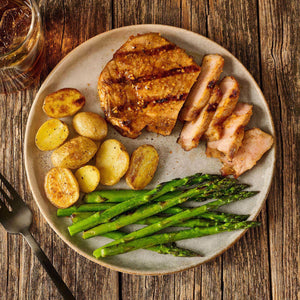 A delicious-looking grilled pork chop, seasoned with Mccormick Grill Mates Bourbon &amp; Oak Seasoning Ground 3.37 Oz, is plated with roasted potatoes and fresh asparagus on a rustic wooden table.