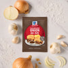 A packet of McCormick Caramelized Onion Au Jus mix is surrounded by onions and garlic.