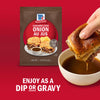 A hand dips a French dip sandwich into au jus gravy next to a packet of McCormick French Onion Au Jus mix.