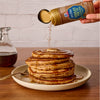 A hand sprinkles Mccormick Harry Potter Chocolate Frog Finishing Sugar, 3.47 Oz over a tall stack of pancakes drizzled with syrup.
