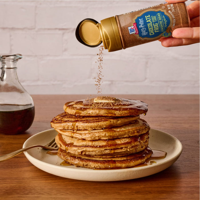A hand sprinkles Mccormick Harry Potter Chocolate Frog Finishing Sugar, 3.47 Oz over a tall stack of pancakes drizzled with syrup.