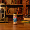 A bottle of Mccormick Harry Potter Chocolate Frog Finishing Sugar, 3.47 Oz, sits on a wooden desk in a cozy library setting next to a French press coffee maker and a stack of books.
