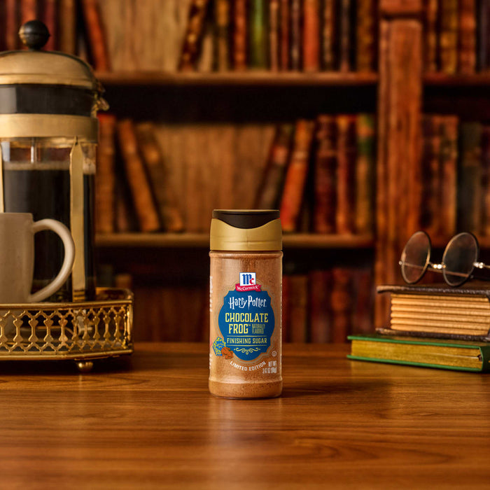 A bottle of Mccormick Harry Potter Chocolate Frog Finishing Sugar, 3.47 Oz, sits on a wooden desk in a cozy library setting next to a French press coffee maker and a stack of books.