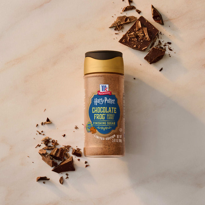 A bottle of Mccormick Harry Potter Chocolate Frog Finishing Sugar, 3.47 Oz is displayed on a marble surface, surrounded by pieces of broken chocolate.