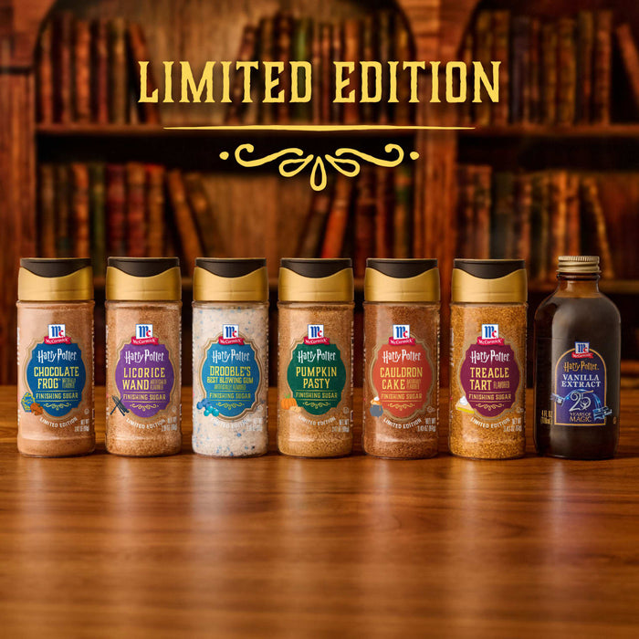 A lineup of limited edition McCormick Harry Potter themed baking products, including a bottle of Mccormick Harry Potter Chocolate Frog Finishing Sugar, 3.47 Oz, is displayed on a wooden table in front of a bookshelf.