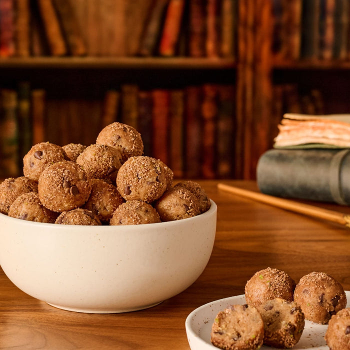 A bowl of chocolate chip cookie dough bites are topped with the coarse, sparkling Mccormick Harry Potter Chocolate Frog Finishing Sugar, 3.47 Oz, and set on a wooden desk in a library.