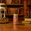 A bottle of Mccormick Harry Potter Cauldron Cake Finishing Sugar, 3.43 Oz is staged on a wooden desk in a library setting, complete with a French press, coffee mug, and a stack of books with glasses, creating a magical and cozy atmosphere.