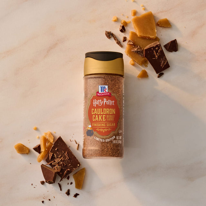 A bottle of Mccormick Harry Potter Cauldron Cake Finishing Sugar, 3.43 Oz is shown on a marble surface surrounded by pieces of chocolate and toffee.