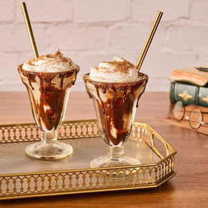 Two decadent chocolate sundaes in classic glasses are presented on a golden tray, each topped with whipped cream and a magical sprinkle of Mccormick Harry Potter Cauldron Cake Finishing Sugar, 3.43 Oz.