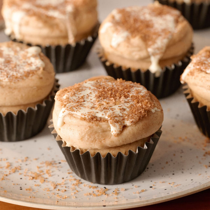 A close-up of several frosted cupcakes in dark liners, beautifully finished with a white glaze and a sprinkle of Mccormick Harry Potter Cauldron Cake Finishing Sugar, 3.43 Oz.