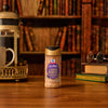 A bottle of Mccormick Harry Potter Licorice Wand Finishing Sugar, 3.39 Oz, sits on a wooden desk in a library setting, accompanied by a coffee press and a stack of books with glasses on top.