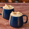 Two blue mugs filled with whipped cream and sprinkled with brown sugar sit on a wooden surface.