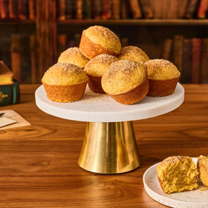 A pile of golden pumpkin pasty muffins, topped with Mccormick Harry Potter Pumpkin Pasty Finishing Sugar, 3.47 Oz, are arranged on a white and gold cake stand in a cozy library setting.