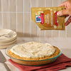A hand sprinkles Mccormick Salted Caramel Cheesecake Seasoning Mix, 1.62 Oz over a freshly prepared cheesecake in a glass dish to add a sweet and salty finish.