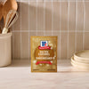 A package of Mccormick Salted Caramel Cheesecake Seasoning Mix, 1.62 Oz stands on a marble countertop in a kitchen setting with wooden utensils and plates.