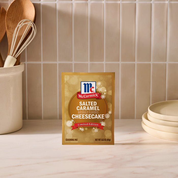 A package of Mccormick Salted Caramel Cheesecake Seasoning Mix, 1.62 Oz stands on a marble countertop in a kitchen setting with wooden utensils and plates.
