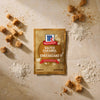 A package of Mccormick Salted Caramel Cheesecake Seasoning Mix, 1.62 Oz is centered on a textured surface surrounded by artistic piles of sea salt flakes and brown sugar cubes.