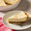 A slice of creamy dessert prepared with Mccormick Salted Caramel Cheesecake Seasoning Mix 1.62 Oz sits on a white plate next to a fork and a red checkered napkin.