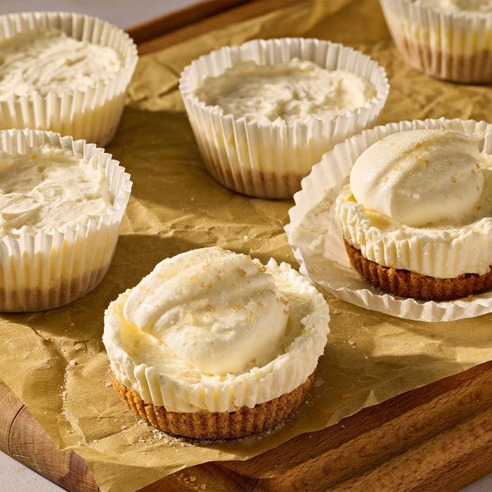Miniature cheesecakes with a graham cracker crust and creamy topping are prepared using Mccormick Salted Caramel Cheesecake Seasoning Mix 1.62 Oz on a parchment lined wooden board.