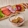 A hand pours a packet of Mccormick Turkey Rub, 1.25 Oz over a bowl of ground meat next to a cutting board with sliced tomatoes and onions.