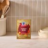 A packet of Mccormick Turkey Rub, 1.25 Oz stands on a marble countertop in a kitchen setting with wooden utensils and stacked plates in the background.