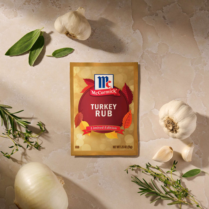 A package of Mccormick Turkey Rub, 1.25 Oz is displayed on a stone surface surrounded by fresh ingredients like whole garlic bulbs, a white onion, and sprigs of rosemary, sage, and thyme.