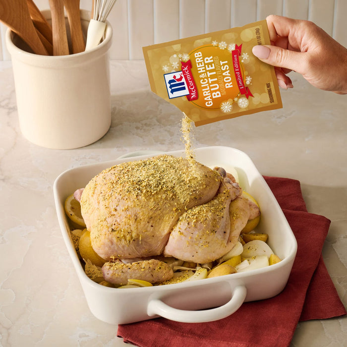 A hand pours a packet of Mccormick Garlic And Herb Butter Roast Seasoning Mix, 1.0 Oz over a whole raw chicken and vegetables in a white roasting dish.