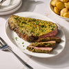 A succulent beef roast is sliced on a white platter and coated in a savory crust made with Mccormick Garlic And Herb Butter Roast Seasoning Mix 1.0 Oz alongside a bowl of roasted potatoes.