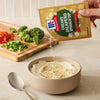 A hand pours a packet of Mccormick Creamy Jalapeno Spinach Dip Seasoning Mix, 0.87 Oz into a bowl of creamy base next to a cutting board with fresh chopped vegetables.
