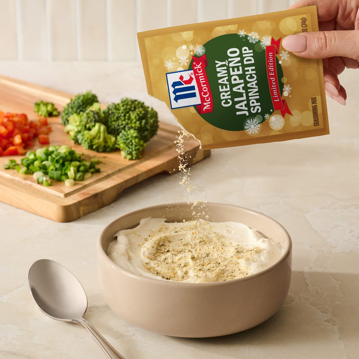A hand pours a packet of Mccormick Creamy Jalapeno Spinach Dip Seasoning Mix, 0.87 Oz into a bowl of creamy base next to a cutting board with fresh chopped vegetables.
