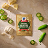 A package of Mccormick Creamy Jalapeno Spinach Dip Seasoning Mix, 0.87 Oz is displayed on a stone surface surrounded by fresh ingredients like whole garlic, sliced jalapenos, and chunks of white cheese.