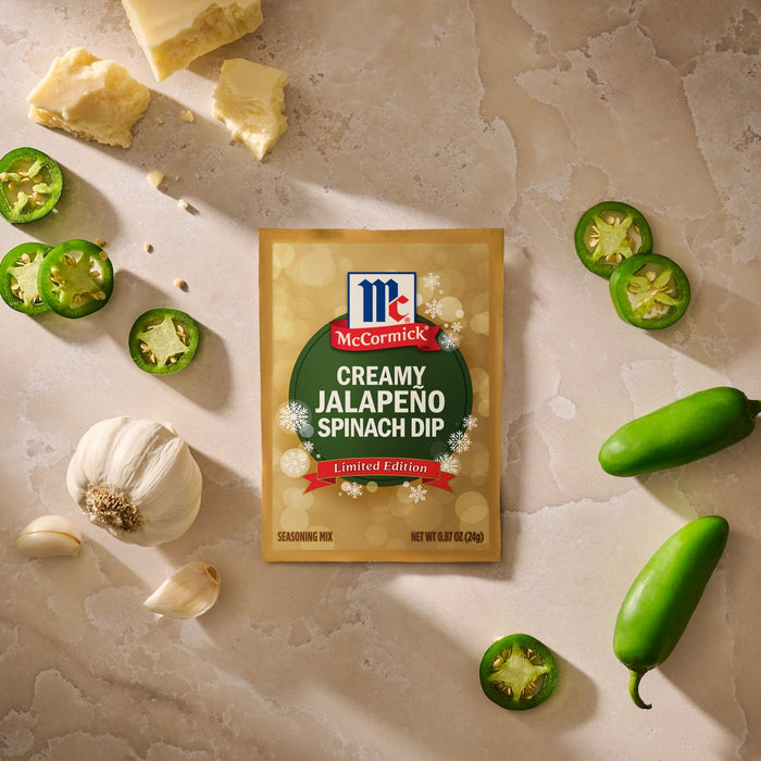 A package of Mccormick Creamy Jalapeno Spinach Dip Seasoning Mix, 0.87 Oz is displayed on a stone surface surrounded by fresh ingredients like whole garlic, sliced jalapenos, and chunks of white cheese.