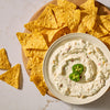 A white bowl filled with a thick dip prepared using Mccormick Creamy Jalapeno Spinach Dip Seasoning Mix 0.87 Oz is garnished with fresh jalapeno slices and served on a wooden platter with yellow tortilla chips.
