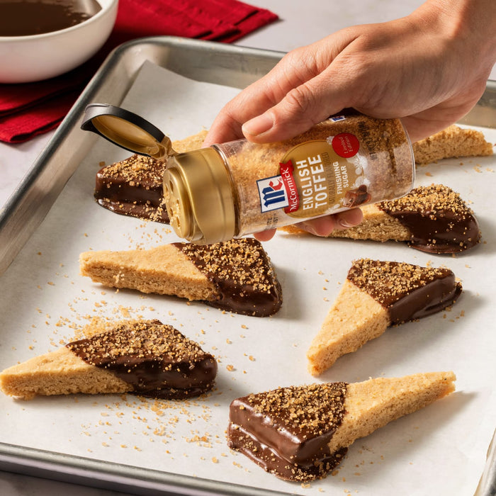A hand sprinkles Mccormick English Toffee Finishing Sgr 3.7 Oz onto a pan of glazed cinnamon rolls.