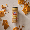 A bottle of Mccormick English Toffee Finishing Sgr 3.7 Oz stands upright amidst scattered pieces of chocolate-covered toffee with almonds and loose toffee bits on a white background.