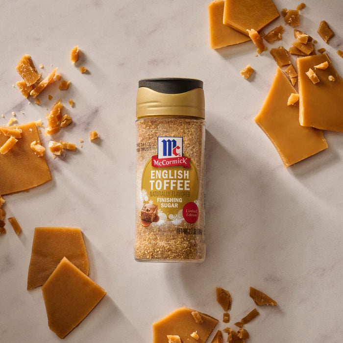 A bottle of Mccormick English Toffee Finishing Sgr 3.7 Oz stands upright amidst scattered pieces of chocolate-covered toffee with almonds and loose toffee bits on a white background.
