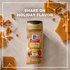 A hand shakes Mccormick English Toffee Finishing Sgr 3.7 Oz onto a pan of glazed cinnamon rolls, adding seasonal flavor.