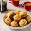 A hand dips a warm muffin into a dish of Mccormick English Toffee Finishing Sgr 3.7 Oz, with other finished muffins on a plate and more baking in a muffin pan in the background.