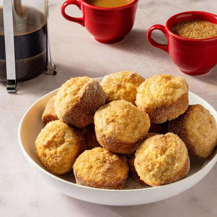 A hand dips a warm muffin into a dish of Mccormick English Toffee Finishing Sgr 3.7 Oz, with other finished muffins on a plate and more baking in a muffin pan in the background.