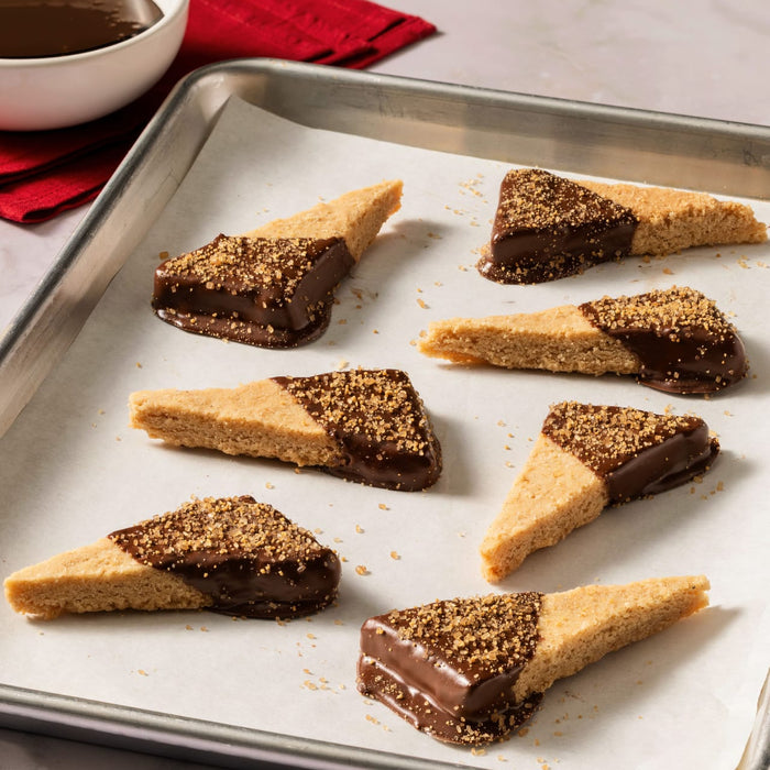 Triangular shortbread cookies, half-dipped in chocolate and sprinkled with finishing sugar, are arranged on parchment paper with a bottle of Mccormick English Toffee Finishing Sgr 3.7 Oz and a bowl of melted chocolate in the background.