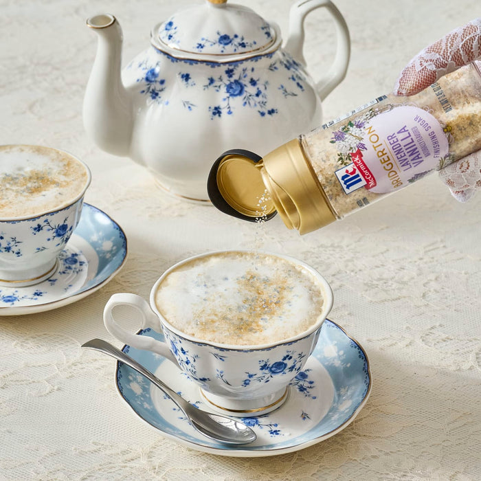 A person sprinkles McCormick Lavender Vanilla Flavored Sugar on a frothy drink served in a blue floral teacup, with a matching teapot and saucer nearby.