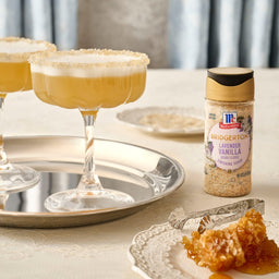 Two cocktails with a sugared rim are displayed next to a McCormick Vanilla Sugar bottle and a plate of honeycomb.