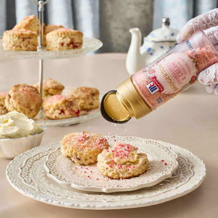 A hand sprinkles McCormick Pink Peppercorn Finishing Salt on scones served with cream on a tiered stand.