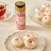 A bottle of McCormick Strawberry Cream Pie Dessert Topping Sprinkles is displayed next to a plate of mini donuts with white frosting and pink sprinkles, accompanied by a cup of tea.
