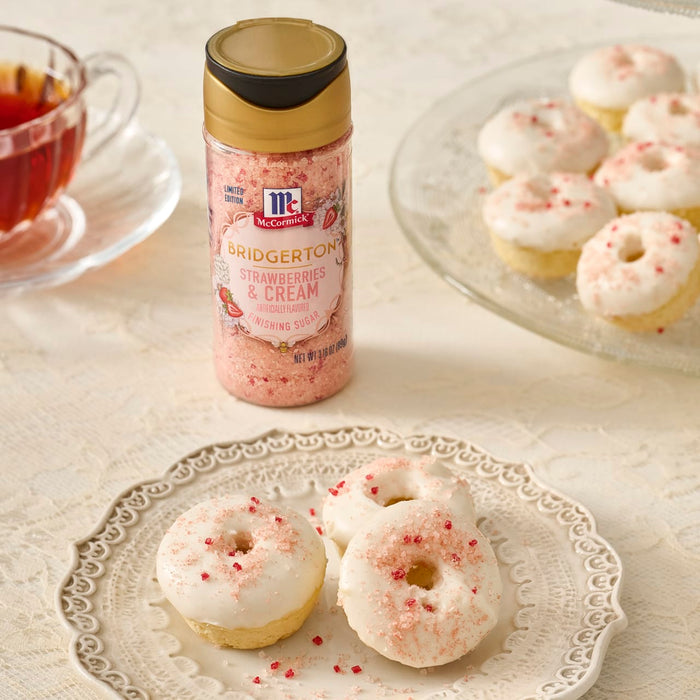 A bottle of McCormick Strawberry Cream Pie Dessert Topping Sprinkles is displayed next to a plate of mini donuts with white frosting and pink sprinkles, accompanied by a cup of tea.