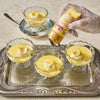 A hand in a lace glove pours a golden sauce from a bottle onto individual servings of custard topped with whipped cream and citrus zest, likely a dessert offering from the brand featured on the bottle.